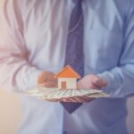 A man in a light blue long-sleeved shirt holding several $100 bills and a figure of a home on top.